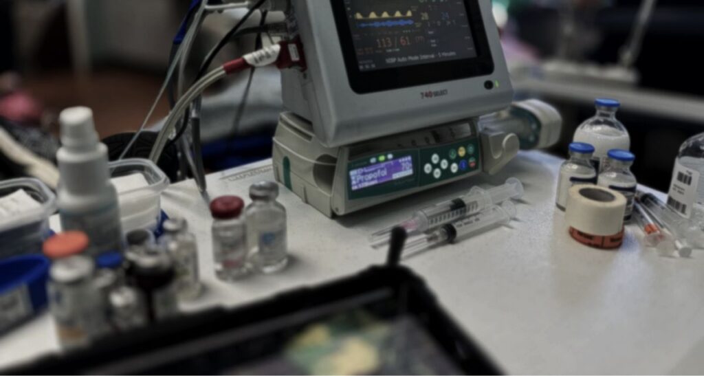 Anesthesia, medical equipment and supplies on a table, including a patient monitor displaying vital signs, vials of medication, syringes, and adhesive tape, in a clinical setting